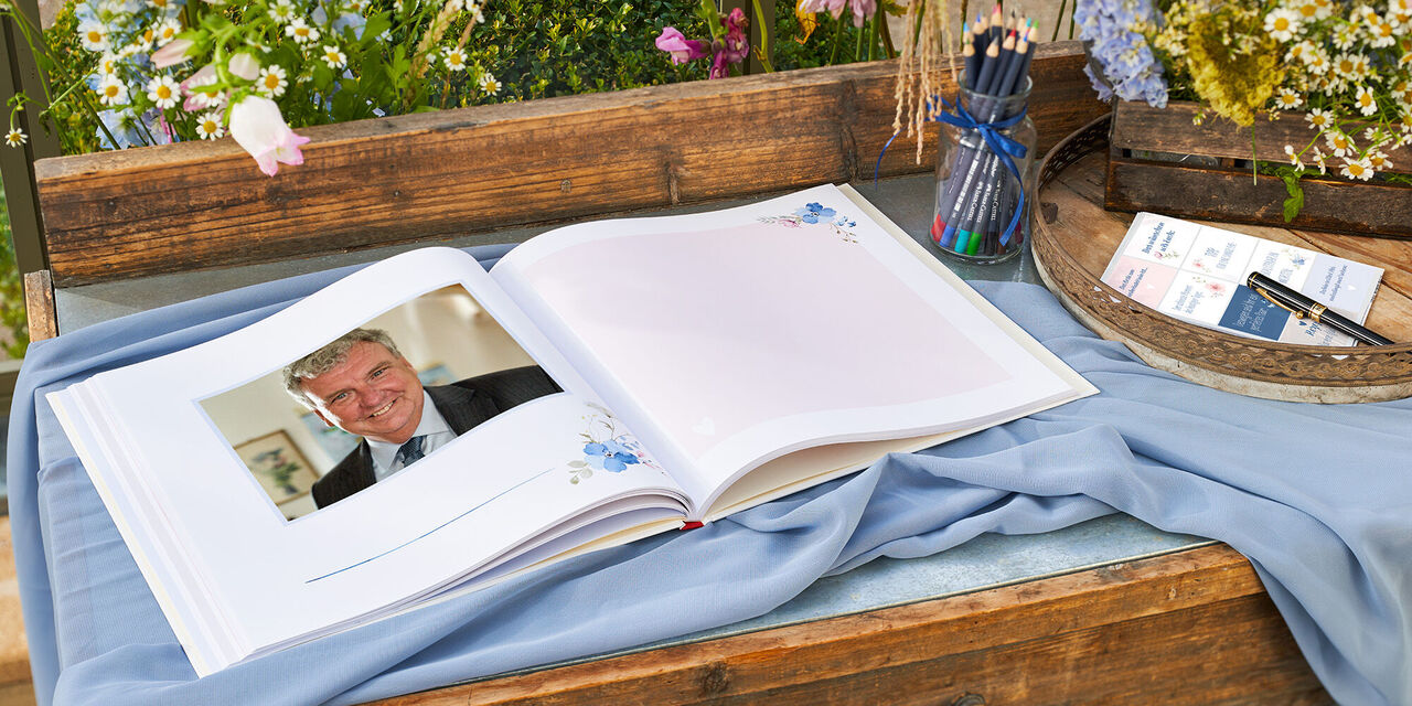 Ein Gästebuch liegt aufgeklappt auf einem Holztisch. Die Doppelseite zeigt links das Foto eines Mannes und rechts einen handschriftlichen Eintrag. Neben dem Buch steht ein Glas mit Stiften. Daneben liegt ein Bogen mit Stickern. Im Hintergrund stehen Blumen.