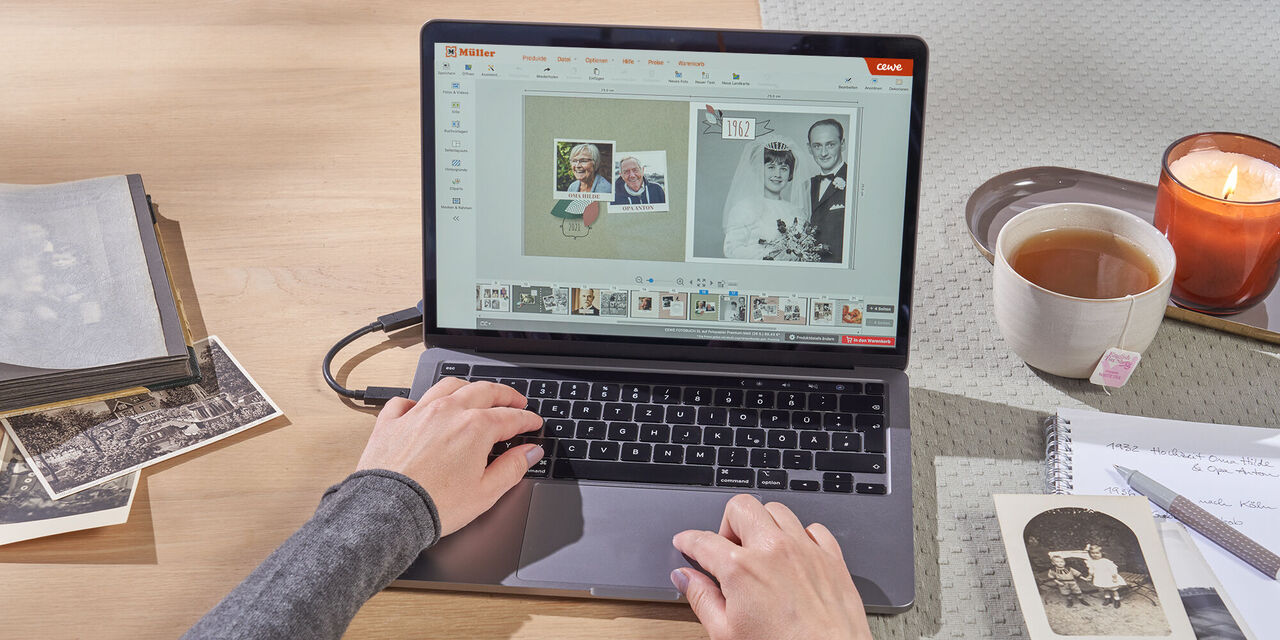 Ein Laptop steht auf einem Tisch. Auf dem Display ist die CEWE Fotowelt Software geöffnet, in der eine Person ein Fotobuch mit der Familienchronik-Buchvorlage bearbeitet. Links neben dem Laptop liegen analoge Schwarzweiß-Fotos. Rechts stehen eine Tasse Tee und eine Kerze.