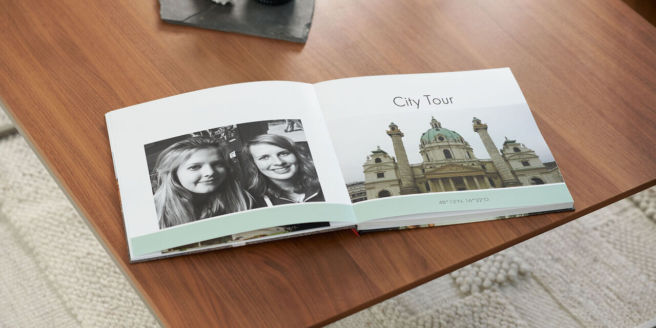 Auf einem Holztisch liegt ein aufgeschlagenes Fotobuch mit einem Schwarz-Weiß-Bild von zwei Frauen und einem Bild einer Kirche sowie der Überschrift Wien. Über dem Fotobuch steht ein dunkles Tablett mit einer Kerze und Muschel. Unter dem Tisch sieht man einen hellen Teppich.