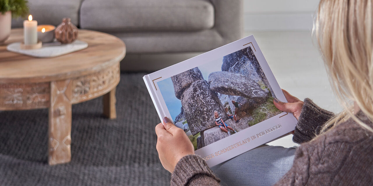 Eine blondhaarige Frau hält ein CEWE FOTOBUCH in den Händen. Auf dem Cover sind Fotos von einer Familie beim Wandern in den Bergen zu sehen.