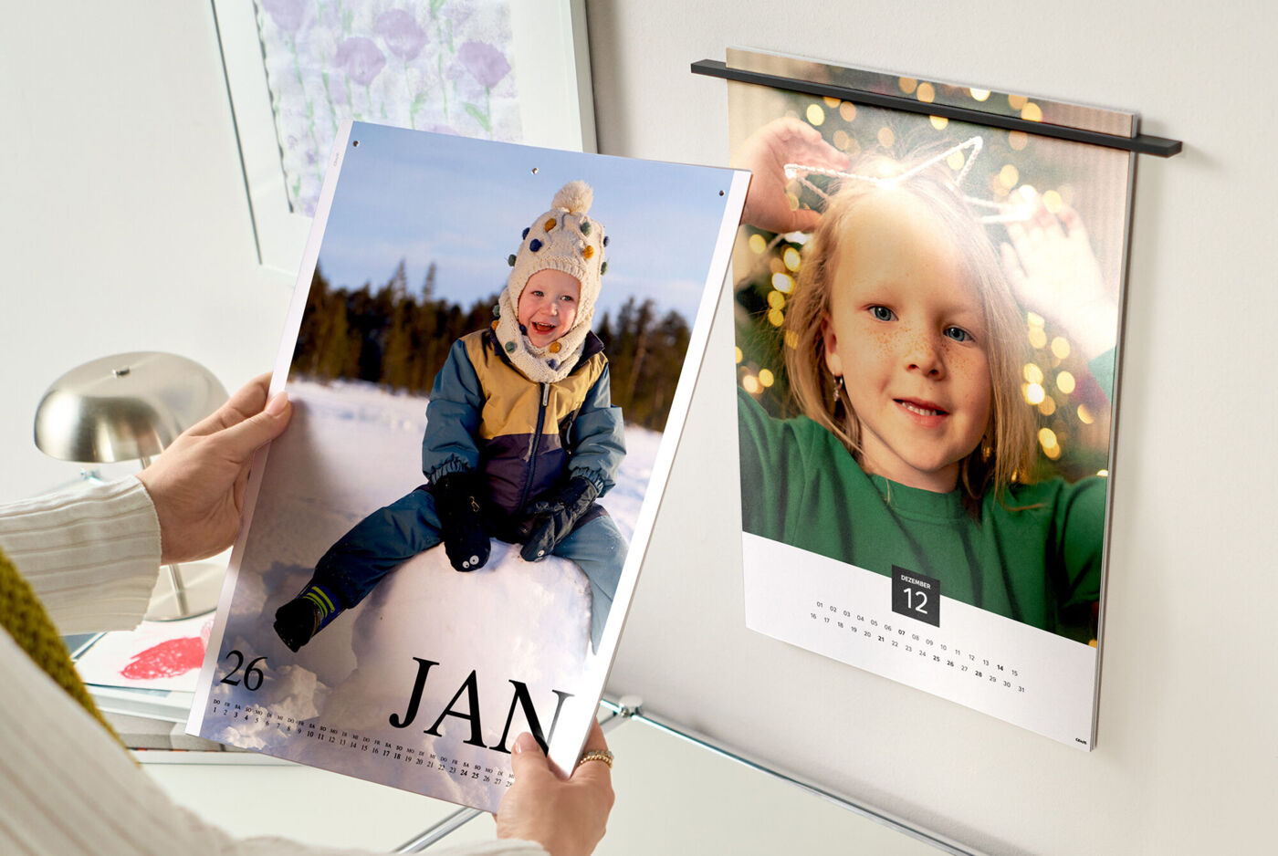 Zwei CEWE Fotokalender: in der Hand Januar mit Kind im Schnee, an der Wand Dezember mit Lichtern