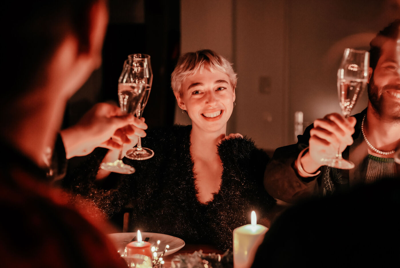 Porträt einer jungen Frau, die im Kerzenschein mit Freunden an einem winterlich geschmückten Tisch sitzt. Gemeinsam heben sie ein Glas Sekt zum anstoßen. Sie lächelt ihr Gegenüber freundlich an.